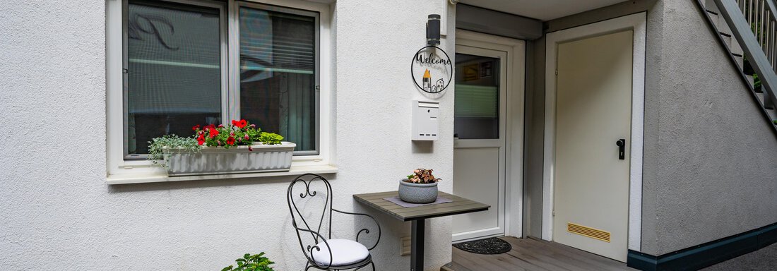An entrance area with a white wall and a small table with a chair. Flowers are on the windowsill and there is a staircase in the background. - Apart Conny - Längenfeld