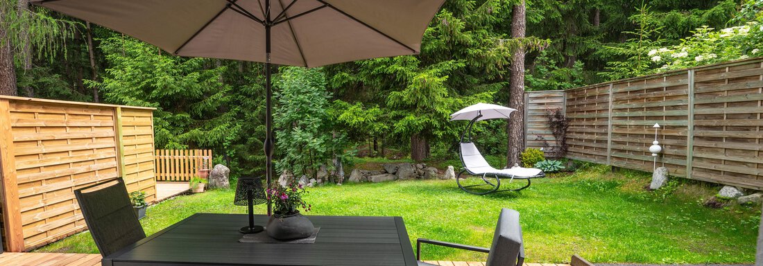 A cozy outdoor area with a table and chairs under a sun umbrella. Surrounded by green trees and lawn, the space offers a relaxed atmosphere. - Apart Conny - Längenfeld