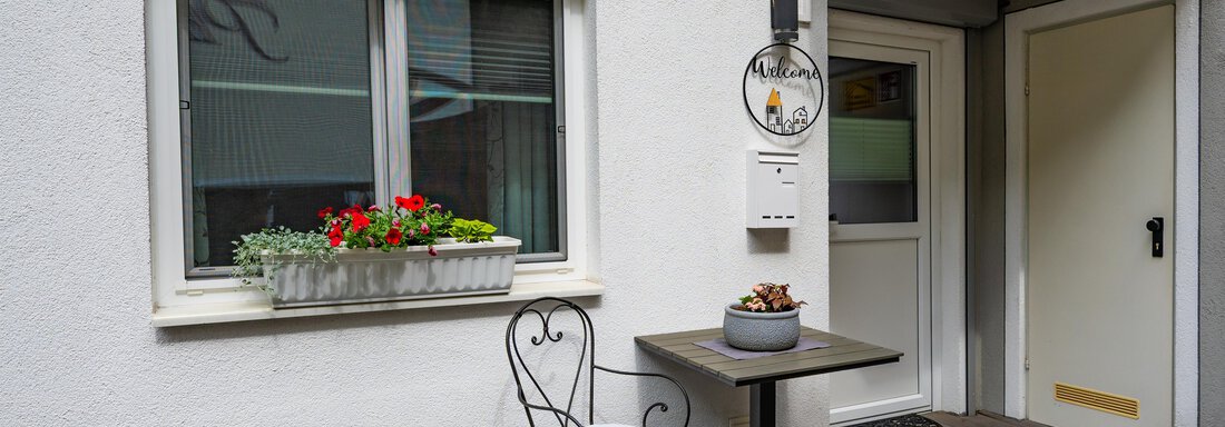 A cozy outdoor area with a table and a chair in front of a white building. Bright flowers in a window box and a green plant create an inviting atmosphere. - Apart Conny - Längenfeld