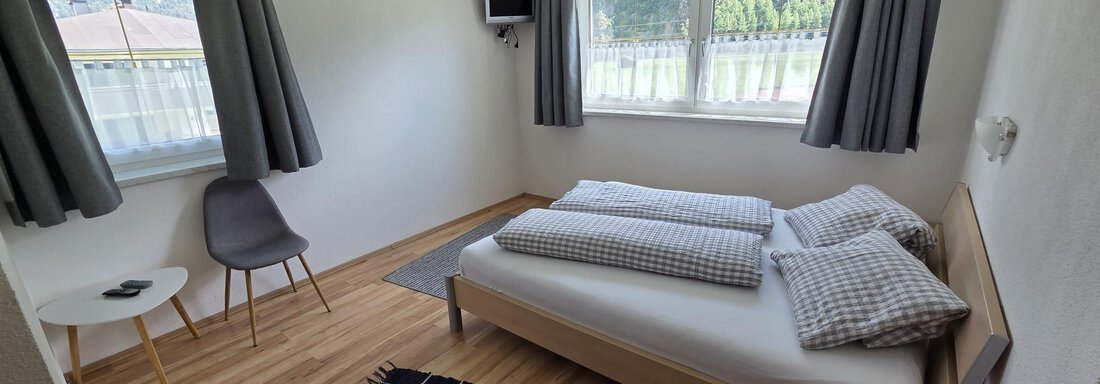 Ein einfaches Schlafzimmer mit einem Bett, zwei Fenstern und einem kleinen Tisch. Der Boden ist aus Holz und ein Teppich liegt auf dem Boden. - Apart Luca - Längenfeld