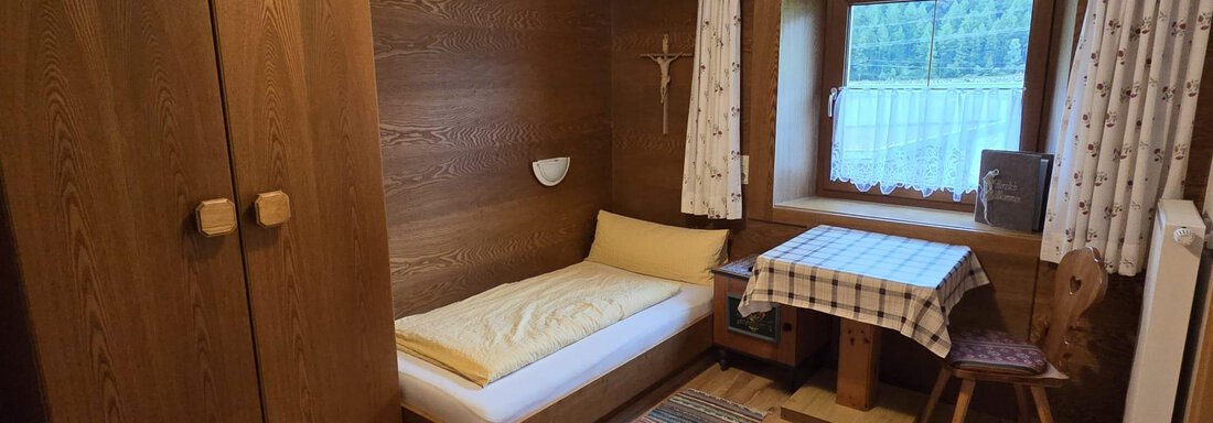 Ein kleines, gemütliches Zimmer mit Holzvertäfelung. Es enthält ein Einzelbett, einen Tisch mit Stuhl und einen Kleiderschrank. - Apart Luca - Längenfeld