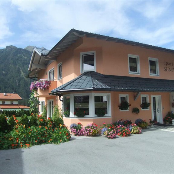 Ein gemütliches, pinkfarbenes Apartmenthaus mit Blumenpflanzen vor der Fassade. Im Hintergrund sind Berge und ein klarer Himmel zu sehen. - Apart Sunshine - Längenfeld