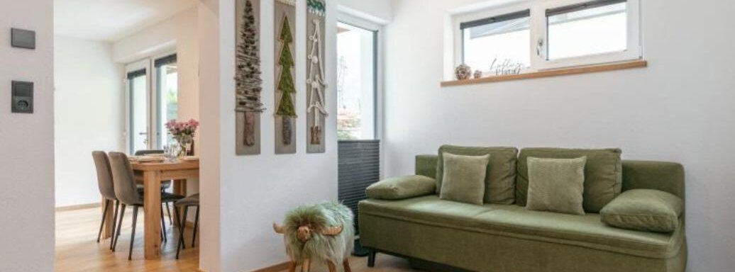 A modern living space with a green sofa and light walls. On the left is a dining table with chairs and decorative elements on the wall. - Apartment DoLa - Längenfeld