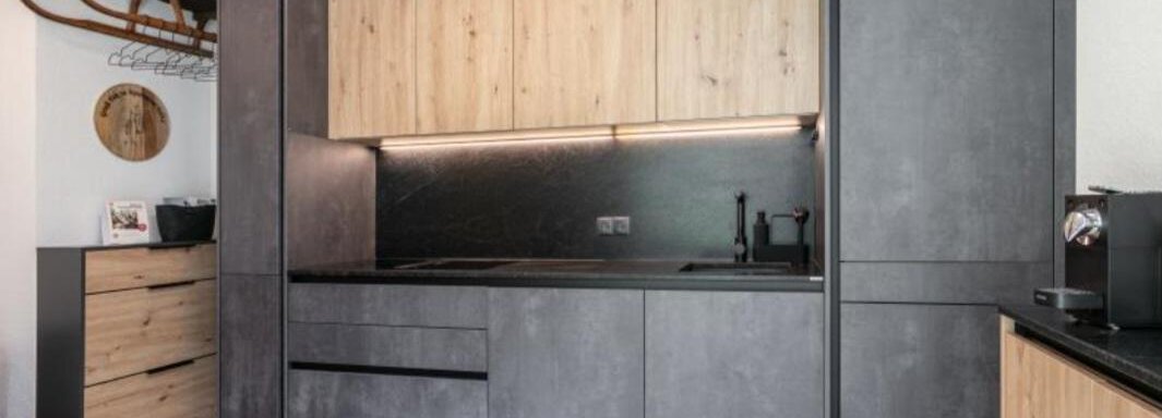 A modern kitchen with black and wood-accented cabinets. The floor is made of light wood and the design is minimalist. - Apartment DoLa - Längenfeld