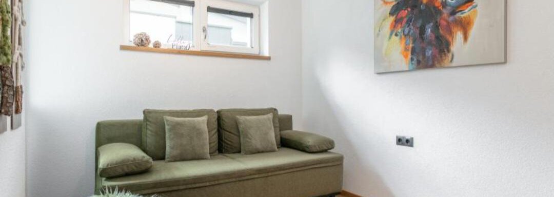 A modern room with a green sofa and a colorful painting on the wall. The floor is made of wood, and there is a small, decorative stool. - Apartment DoLa - Längenfeld