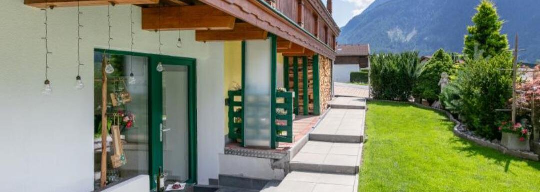 Ein modernes Haus mit einer Terrasse und einem gepflegten Garten. Im Hintergrund sind die Berge und ein klarer Himmel zu sehen. - Apartment DoLa - Längenfeld