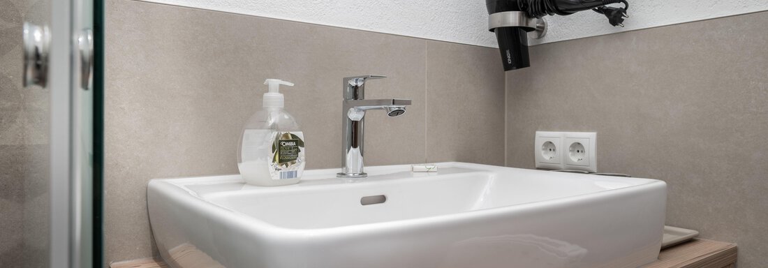 Ein modernes Badezimmer mit einem rechteckigen Waschbecken auf einer Holzablage. Im Hintergrund sind ein Haartrockner und Pflegeprodukte zu sehen. - Appart Klotz Alpenglück - Längenfeld