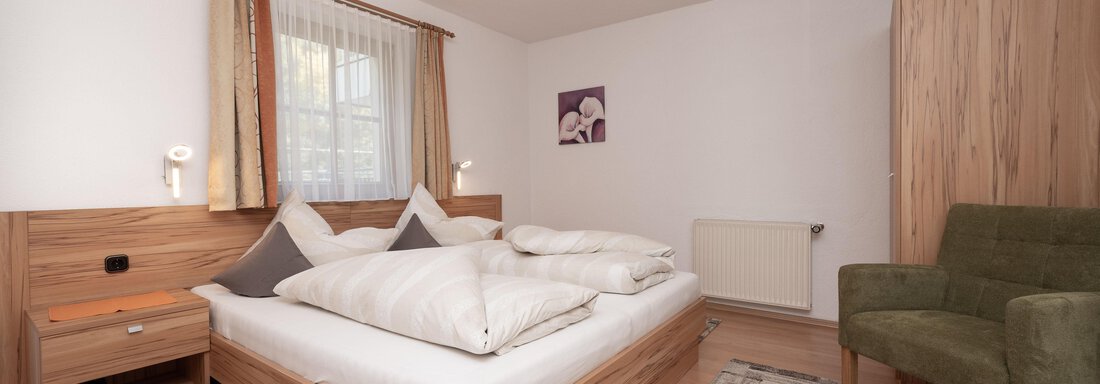 Ein gemütliches Schlafzimmer mit einem Doppelbett und frisch bezogenen Kissen. Neben dem Bett steht ein gemütlicher Sessel und eine Holzkommode. - Appart Klotz Alpenglück - Längenfeld