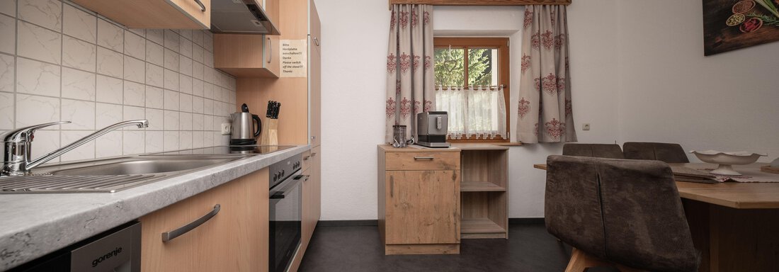 Eine moderne Küche mit Holzschränken und grauer Arbeitsplatte. Der Raum ist hell und einladend, mit einem Essbereich und Fenstern mit Vorhängen. - Appart Klotz Alpenglück - Längenfeld