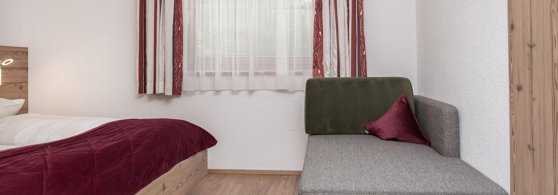Ein gemütliches Zimmer mit einem Doppelbett und einem Grauen Sofa. Die Fenster sind mit hellen Vorhängen geschmückt, und der Boden ist aus Holz. - Appart Klotz Alpenglück - Längenfeld