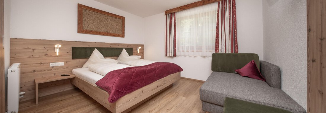 Ein modernes Zimmer mit einem Doppelbett, grauem Sofa und warmen Holzdetails. Große Fenster sorgen für viel Tageslicht. - Appart Klotz Alpenglück - Längenfeld