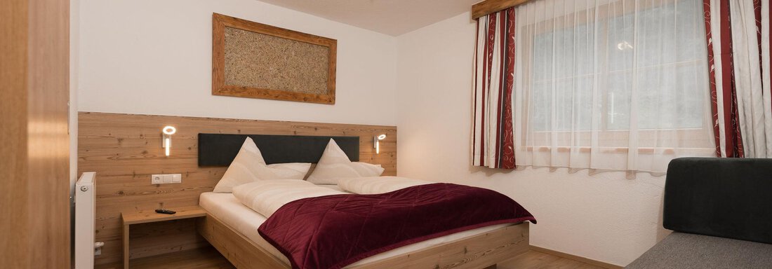 Ein modernes Schlafzimmer mit einem Doppelbett und roten Akzenten. Es gibt einen gemütlichen Sessel und große Fenster, die viel Licht hereinlassen. - Appart Klotz Alpenglück - Längenfeld