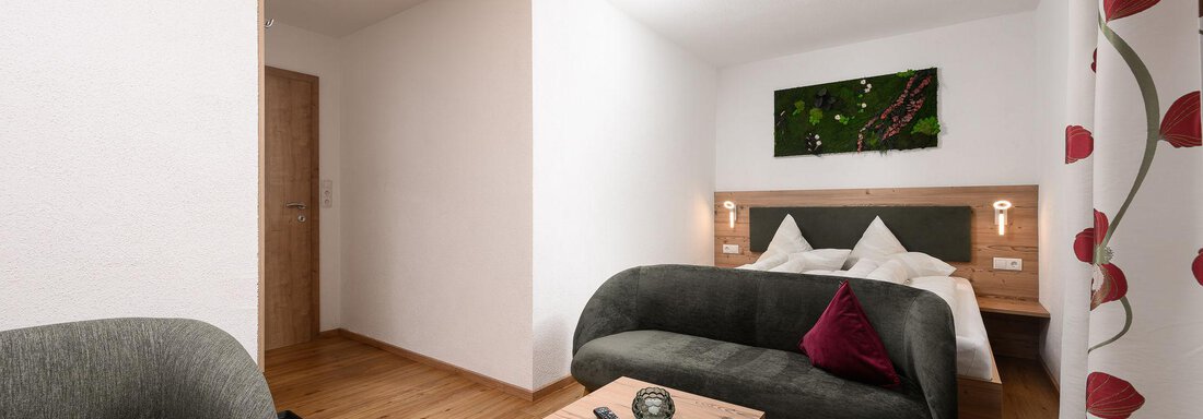 Ein modernes Zimmer mit Holzfußboden und hellen Wänden. Es gibt ein bequemes Sofa, einen kleinen Tisch und ein großes Bett im Hintergrund. - Appart Klotz Alpenglück - Längenfeld