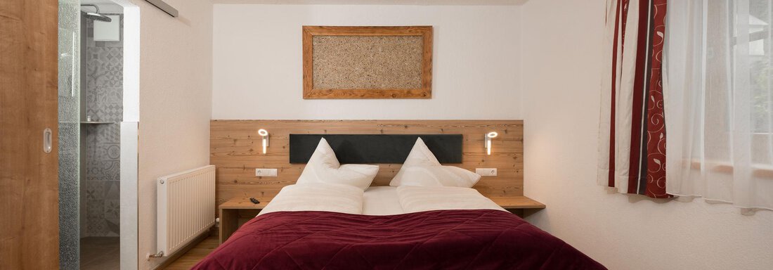Ein gemütliches Schlafzimmer mit einem großen Bett und rot gemusterten Kissen. Die Wände sind hell, und es gibt einen modernen Badezimmerbereich im Hintergrund. - Appart Klotz Alpenglück - Längenfeld
