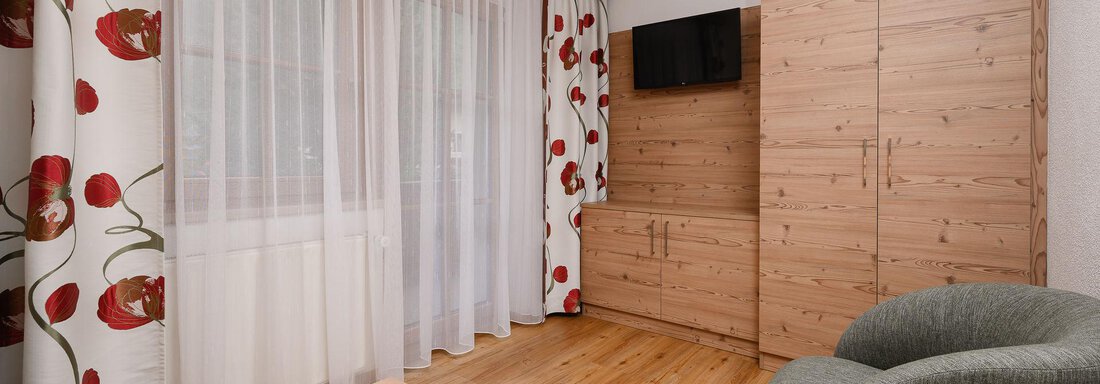 Ein helles Zimmer mit Holzfußboden und großen Fenstern. Die Wände sind mit Vorhängen in Blumenmuster dekoriert, und es gibt eine graue Sitzgelegenheit sowie einen Fernseher. - Appart Klotz Alpenglück - Längenfeld