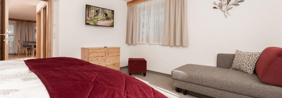 Ein gemütliches Schlafzimmer mit einem großen Bett und einem grauen Sofa. An der Wand hängt ein Fernseher und es gibt helle Vorhänge, die Licht hereinlassen. - Appart Klotz Alpenglück - Längenfeld