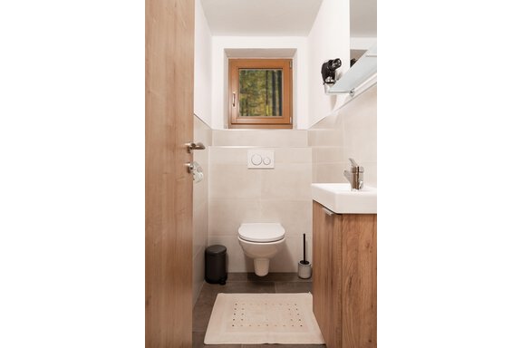 Ein modernes Badezimmer mit einer Toilette und einem Waschbecken. Ein Fenster lässt natürliches Licht herein und zeigt einen grünen Ausblick. - Appart Klotz Alpenglück - Längenfeld