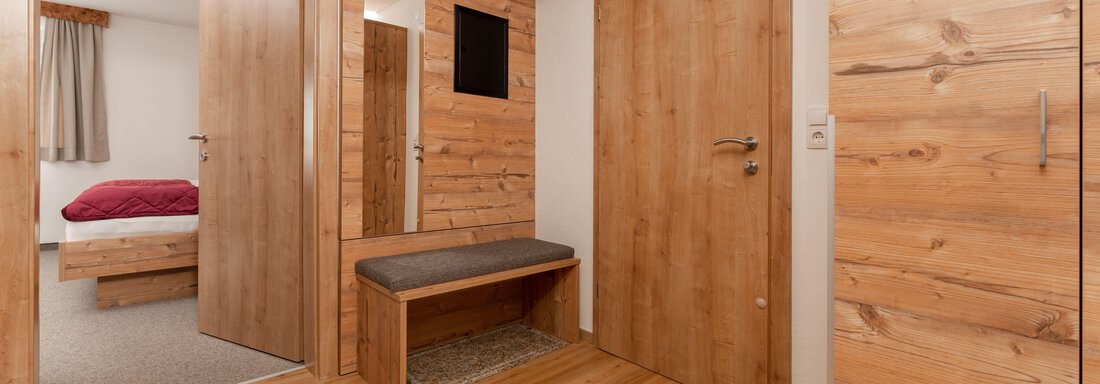 Ein heller Eingangsbereich mit Holzverkleidung und einem Sitzplatz. Im Hintergrund ist ein Schlafzimmer mit einem roten Bett sichtbar. - Appart Klotz Alpenglück - Längenfeld