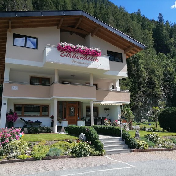 Ein schönes, modernes Haus mit Blumenbalkonen und gepflegtem Garten. Umgeben von grünen Bäumen und einer klaren blauen Himmel. - Birkenheim Widmann - Längenfeld