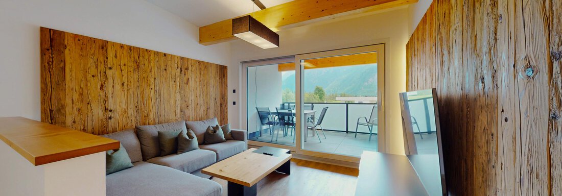 Ein modernes Wohnzimmer mit einer grauen Couch und Holzverkleidungen. Große Fenster bieten einen Blick auf einen Balkon und die umliegende Landschaft. - Das Berg Loft - Längenfeld