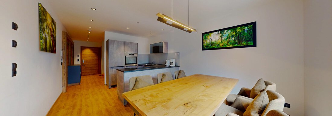 Das Berg Loft Längenfeld - Küche-Esszimmer (2) Ein modernes Wohnzimmer mit einem großen Holztisch und gemütlichen Sitzplätzen. Die Küche ist offen und verfügt über moderne Geräte sowie dekorative Kunstwerke an den Wänden. - Das Berg Loft - Längenfeld