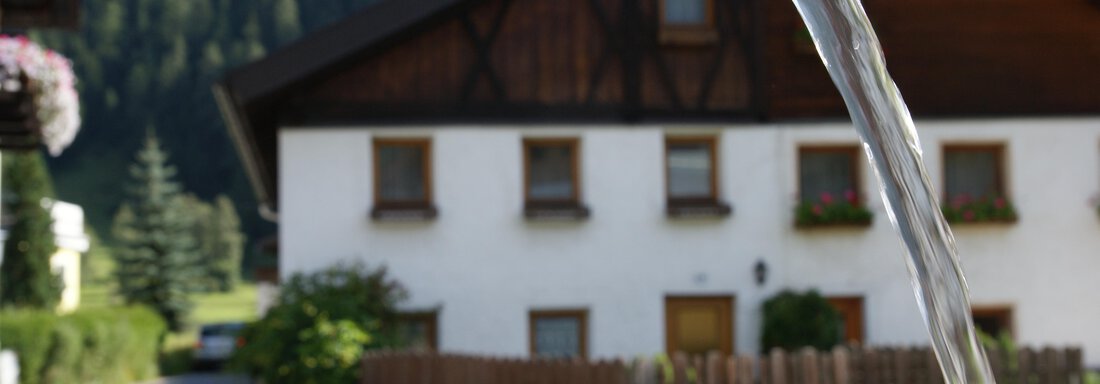 Ein rustikales Haus mit weißen Wänden und brauner Holzverkleidung. Im Vordergrund fließt frisches Wasser aus einem Brunnen. - Ferienhaus Volgger - Längenfeld