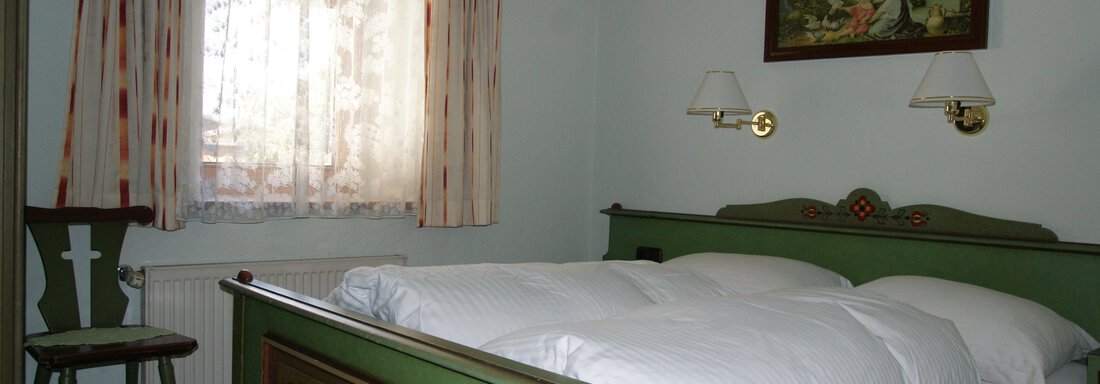 Ein gemütliches Schlafzimmer mit einem großen Bett und einer schönen, bemalten Wanddeko. Ein Fenster mit Vorhängen und ein Stuhl ergänzen die Einrichtung. - Ferienhaus Volgger - Längenfeld