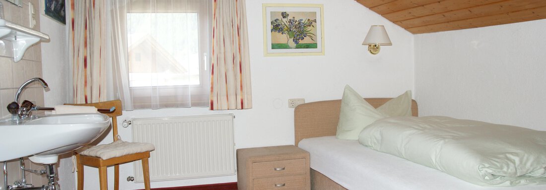 Ein einfaches Zimmer mit einem Einzelbett und einem Fenster. Die Wände sind hell, und es gibt eine kleine Sitzecke sowie einen Waschplatz. - Ferienhaus Volgger - Längenfeld
