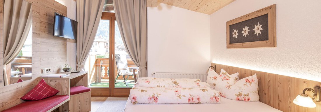 A cozy room with wooden furniture and a large double bed.  
Large windows offer a view of the balcony and let in plenty of light. - Gästehaus Waltraud - Längenfeld