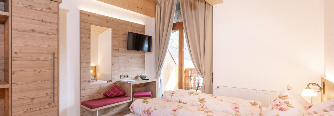 A cozy bedroom with wooden decor and a large bed. There is a mirror, a television, and a balcony with a view outside. - Gästehaus Waltraud - Längenfeld