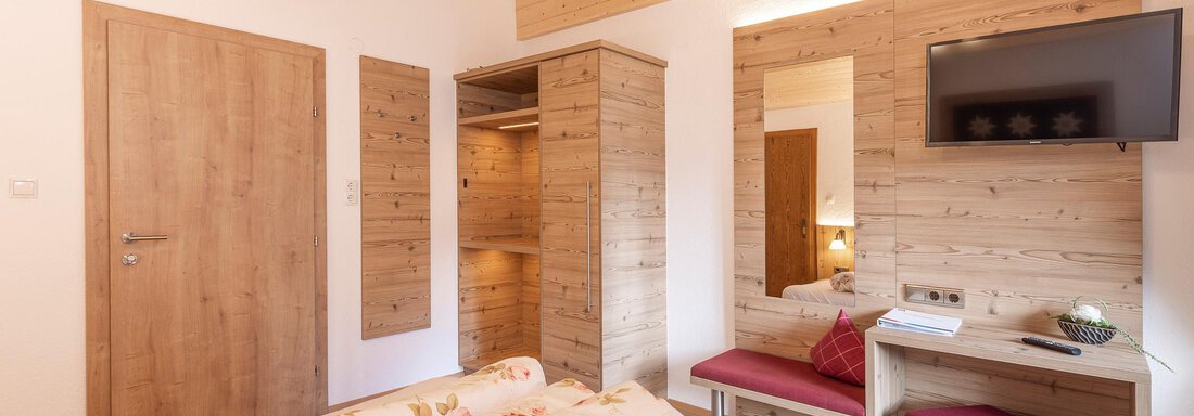 A modern room with wooden interiors and floral bedding. The room is bright and cozy, with a television and a view of nature. - Gästehaus Waltraud - Längenfeld