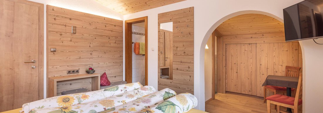 A cozy bedroom with wooden paneling and warm light. There is a large bed with flower bedding and a small table with chairs. - Gästehaus Waltraud - Längenfeld