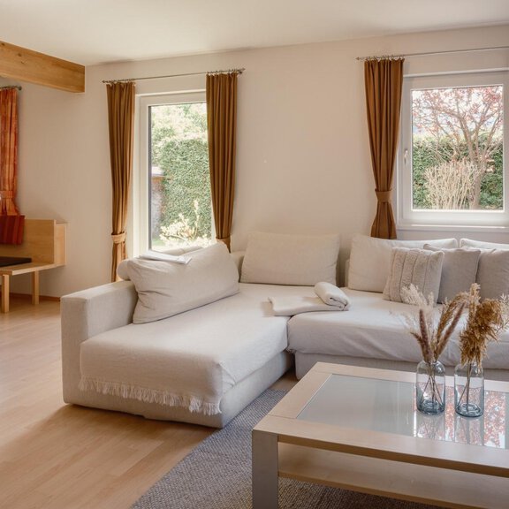 Ein helles Wohnzimmer mit einem bequemen Sofa und einem Glastisch. Durch die Fenster fällt natürliches Licht und eine gemütliche Atmosphäre entsteht. - Haus Margret - Längenfeld