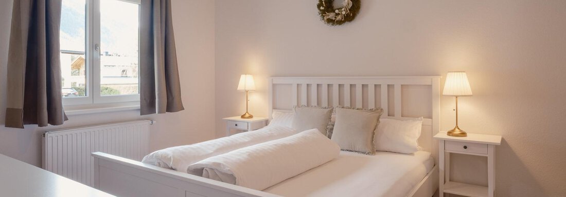 A cozy bedroom with a white bed and soft pillows. The windows let in a lot of light and there are two nightstands with lamps. - Haus Margret - Längenfeld