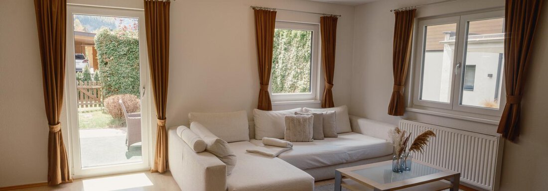 A bright living room with a white sofa and a coffee table. Large windows let in plenty of light and offer a view of the garden. - Haus Margret - Längenfeld