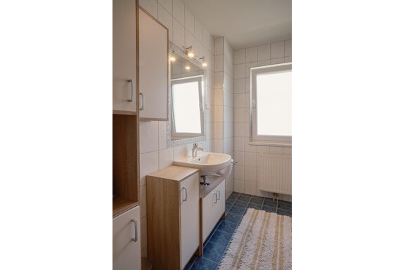 A modern bathroom with white tiles and a large window. It features a sink area with storage and a bright carpet. - Haus Margret - Längenfeld