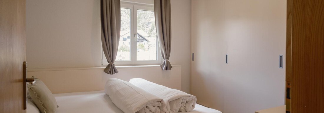 A simple bedroom with a double bed and a large wardrobe. Bright curtains let daylight in. - Haus Margret - Längenfeld