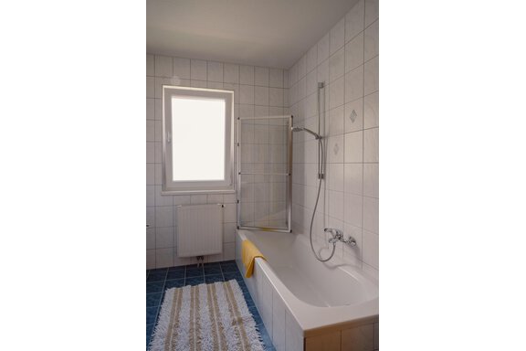A bright bathroom with a bathtub and a shower partition. The walls are covered with white tiles and a window provides daylight. - Haus Margret - Längenfeld