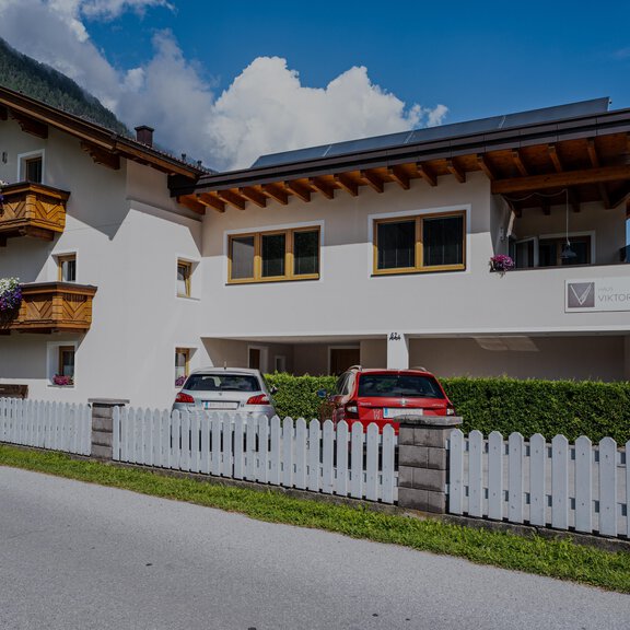 Ein modernes Haus mit ansprechender Holzverkleidung und bunten Balkonblumen. Im Vordergrund befinden sich zwei Autos und ein weißer Zaun. - Haus Viktoria - Längenfeld