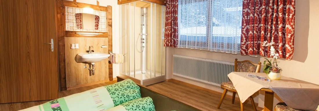 A cozy room with two beds, a small seating area, and a sink. The windows are decorated with bright curtains and the room has a warm, inviting character. - Haus Waldhof - Längenfeld