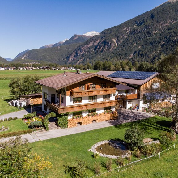 A beautiful multi-story house in the mountains, surrounded by meadows and trees. In the background, majestic mountains and a clear blue sky can be seen. - Hauseggerhof - Längenfeld