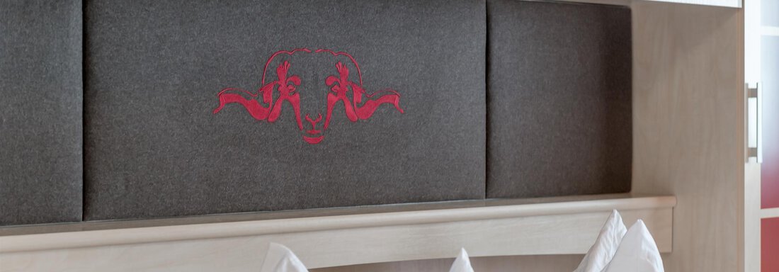 A modern bedroom with a gray headboard and white pillows. The headboard features a red decorative element. - Hotel Burgstein alpin & lifestyle - Längenfeld