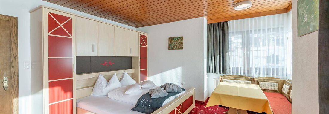A bright room with a double bed and a cozy seating area. The floor is covered with a red carpet, and large windows provide plenty of daylight. - Hotel Burgstein alpin & lifestyle - Längenfeld