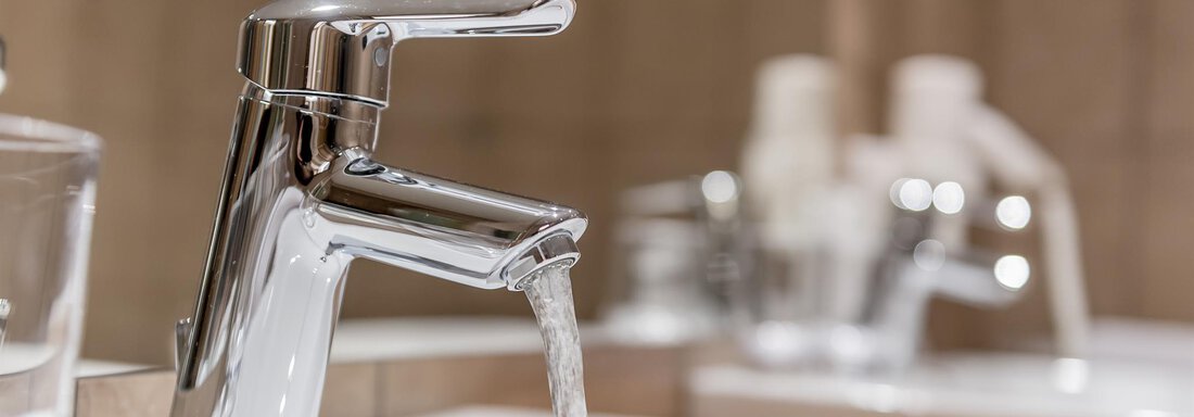 A modern faucet from which clear water flows. In the background, soap dispensers and a glass are visible. - Hotel Burgstein alpin & lifestyle - Längenfeld