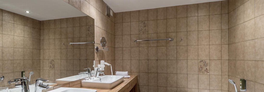 A modern bathroom with two sinks and a bathtub. The walls are designed with light tiles and there are large mirrors over the sinks. - Hotel Burgstein alpin & lifestyle - Längenfeld