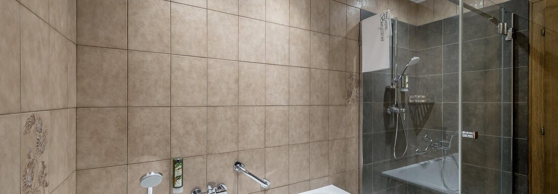A modern bathroom with a bathtub and a separate shower. The walls are tiled with brown tiles and illuminated by several ceiling lights. - Hotel Burgstein alpin & lifestyle - Längenfeld