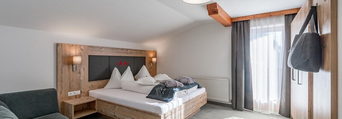 A modern bedroom with wood details and a large window. There is a comfortable bed and a cozy seating area. - Hotel Burgstein alpin & lifestyle - Längenfeld