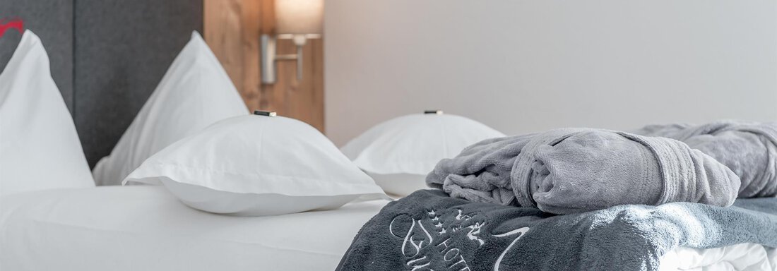 A cozy hotel room with a large bed and white pillows. On it lies a gray towel and a soft bathrobe. - Hotel Burgstein alpin & lifestyle - Längenfeld