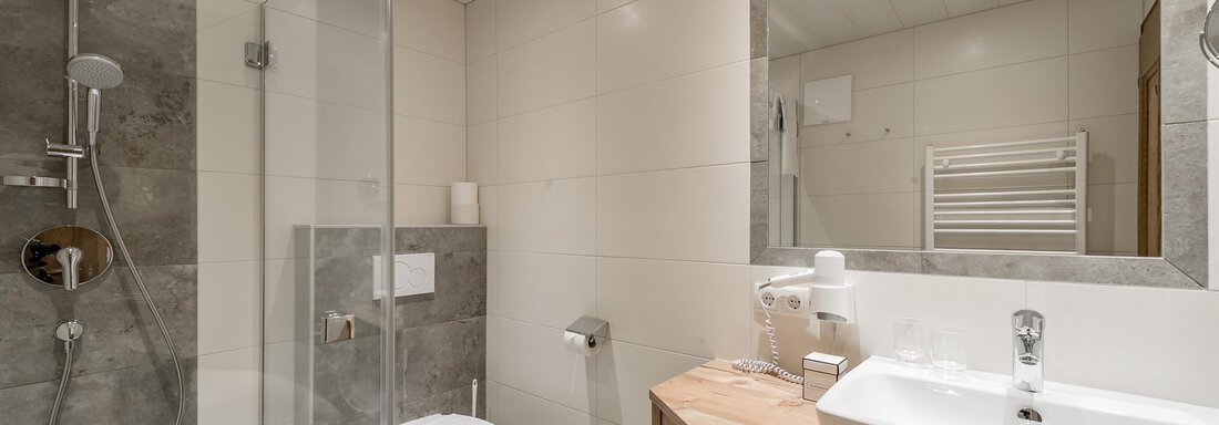 A modern bathroom with a shower stall and an elegant sink. The walls are brightly tiled, and the wooden shelf offers additional storage space. - Hotel Burgstein alpin & lifestyle - Längenfeld