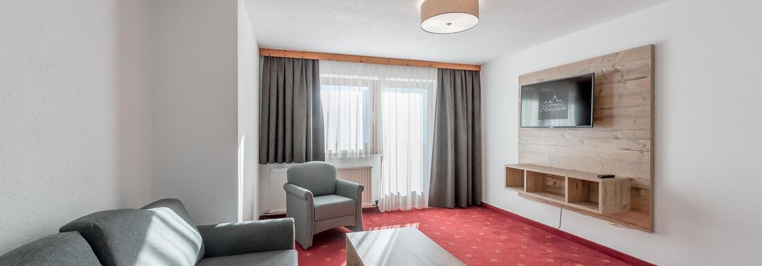 A modern room with a sofa, a table, and a television on the wall. Large windows let in plenty of light, and the furniture is in neutral colors. - Hotel Burgstein alpin & lifestyle - Längenfeld
